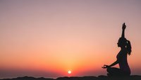 Woman practicing yoga meditation at sunset with peaceful mountain landscape silhouette against orange pink sky for wellness mindfulness spiritual health.
