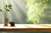 Small green plant in white ceramic pot and tea bowl on wooden table against blurred natural window background with sunlight and foliage.