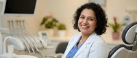 Smiling female dentist with curly dark hair and medium skin tone wearing light blue blazer and blue scrubs in modern dental office clinic setting.