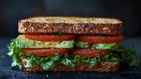 Fresh sandwich with crispy toasted bread, layered with ripe tomato slices, creamy avocado, and crisp lettuce on dark background, close-up view.