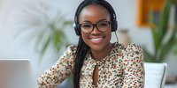 Young African American businesswoman wearing headphones and glasses smiles while working from home office, using laptop for remote work or online meeting.