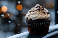 Chocolate cupcake with cream frosting and sprinkles on snowy windowsill with warm bokeh lights creating cozy winter atmosphere for holiday baking and dessert concepts.