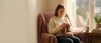 Young Asian woman in cozy cream sweater relaxing in comfortable armchair by sunny window, enjoying warm beverage in peaceful home interior setting.