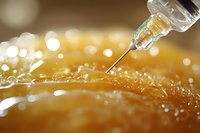 Medical syringe needle injecting liquid medicine into orange textured surface with golden bokeh lights and water drops in macro close up view.