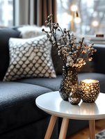 Cozy home interior with dried flowers in dark vase and glowing candle holders on white side table, grey sofa with geometric pattern cushion in background.