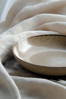 Handcrafted ceramic bowl with speckled glaze on soft white linen fabric, creating gentle folds and shadows in natural light for minimalist food photography styling.