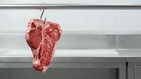 Raw beef steak hanging from hook in modern kitchen with stainless steel shelving, showcasing premium meat quality for culinary and food photography use.