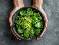 Hands holding bowl of fresh green salad with sliced avocado and spinach leaves on dark stone surface for healthy eating and nutrition concepts.