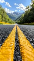 Mountain highway with yellow double lines stretching toward distant peaks, surrounded by lush forest under blue summer sky with white clouds.