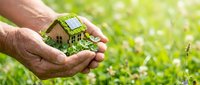 Hands holding miniature eco house with solar panels and green plants against blurred natural background for sustainable living and environmental protection.