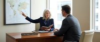 Professional blonde businesswoman presenting global strategy to male colleague in modern corporate office with world map during executive meeting.