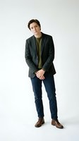 Young man with dark hair and fair complexion wearing charcoal blazer, olive t-shirt, navy jeans and brown leather shoes, standing against white background for fashion and lifestyle use.