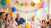 Group of diverse people celebrating at indoor party with colorful balloons and decorations, holding white balloon in foreground, festive gathering for birthday or special event celebration.