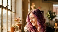 Young woman with purple pink hair smiling by sunny window in cozy home interior with plants and warm natural lighting for lifestyle content.