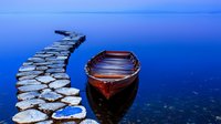 Wooden boat moored near curved stone pathway over tranquil blue water at dusk, creating serene minimalist composition with reflections on calm lake surface.