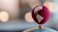 Pink knitted hot air balloon with world map pattern floating above blurred map background with soft bokeh lights creating dreamy travel atmosphere.