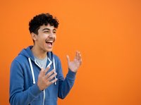Young man with dark curly hair and olive complexion wearing blue hoodie, laughing and gesturing expressively against orange background for lifestyle and emotion content.