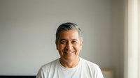 Middle-aged Hispanic man smiling warmly in white t-shirt against neutral wall background for lifestyle and wellness content.