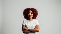 Confident young African American woman with curly red hair smiling with arms crossed wearing white shirt on neutral background for business content.