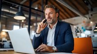 Mature Caucasian businessman in navy suit talking on phone while working on laptop in modern office space with industrial ceiling and natural light.