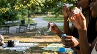 Young diverse women blowing soap bubbles in sunny park during outdoor leisure activity. Friends enjoying summer day with bubble maker toys on green nature background.