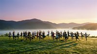 Large group of people standing in circle on grassy field at sunrise near misty lake and mountains, suitable for team building, community, corporate events, and outdoor gathering concepts.