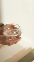 Hand holding clear glass of water against neutral beige background. Male hand, caucasian ethnicity, casual lifestyle setting for hydration, wellness and daily routine concepts.