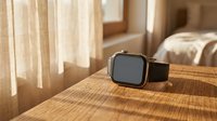 Modern smartwatch with black sport band resting on wooden surface in warm sunlit bedroom interior with natural lighting and soft shadows.