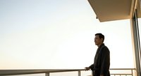 Middle-aged man of Asian ethnicity wearing dark business casual clothing standing on a modern balcony or terrace overlooking a minimalist landscape at golden hour, contemplative mood.