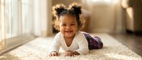 Happy mixed-race toddler girl with curly hair in space buns crawling on carpet in sunny living room, smiling at camera with natural window light.