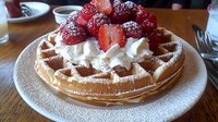 Fresh strawberries and whipped cream top a golden Belgian waffle dusted with powdered sugar, served on white plate against wooden table background.