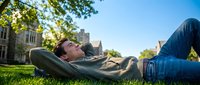 Young man relaxing on green grass in urban park with hands behind head, enjoying sunny day outdoors. Casual lifestyle and leisure concept.
