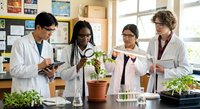 Diverse group of young scientists in white lab coats conducting plant research experiment in bright laboratory with seedlings and scientific equipment.