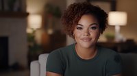 Young African American woman with natural curly hair smiling warmly at camera while sitting in cozy living room. Professional headshot style portrait in casual green t-shirt.