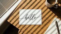 Flat lay of a hello greeting card on a wooden desk with coffee cup and notebook in warm sunlight with shadow stripes.