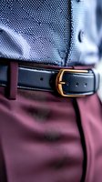Close-up view of black leather belt with gold buckle against burgundy pants and blue shirt, showcasing elegant men's fashion accessories and formal attire details.