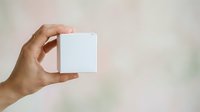 Close-up of hand holding blank white box against neutral background, suitable for product packaging mockups, branding presentations, and commercial advertising campaigns.