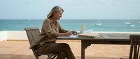 Senior man with gray hair working on laptop at wooden table on coastal terrace overlooking turquoise ocean, remote work and retirement lifestyle concept.