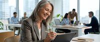 Mature businesswoman with gray hair smiling while using laptop in modern office environment with colleagues working in background.