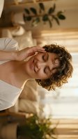 Young woman with curly brown hair wearing white tank top, touching head with hand, smiling with eyes closed in spa or wellness setting with plants and natural light.