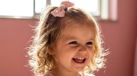 Happy blonde toddler girl with curly hair and pink bow smiling joyfully in bright sunlit room. Cheerful child portrait for family lifestyle content.