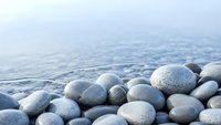 Smooth gray pebbles scattered on sandy beach shore with gentle ocean waves in soft natural lighting for spa wellness and meditation concepts.