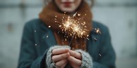 Young woman holding burning sparkler with golden sparks flying, celebrating holidays in cozy winter sweater with warm lighting atmosphere.