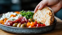 Fresh Mediterranean appetizer plate with prosciutto, cherry tomatoes, olives, and crusty artisan bread served on rustic ceramic platter, hand reaching for bread.