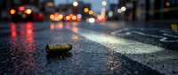 Bullet casing on wet asphalt street at night with blurred city lights and traffic in background creating dramatic crime scene atmosphere.