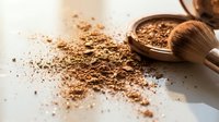 Loose bronze powder scattered on white surface with wooden bowl and soft bristle brush for makeup application and cosmetic artistry.