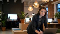 Professional businesswoman working late in modern office with geometric pendant lights and plants creating productive workspace atmosphere.
