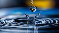 Heart-shaped water droplet floating above crown splash formation in dark blue water with yellow background highlights. Macro photography captures liquid dynamics.
