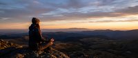 Young woman hiker sitting on mountain peak during golden hour sunset, enjoying panoramic valley view. Adventure travel and outdoor recreation concept.