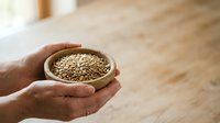 Hand holding wooden bowl filled with sesame seeds on wooden surface. Suitable for food, nutrition, cooking, and healthy eating content.
